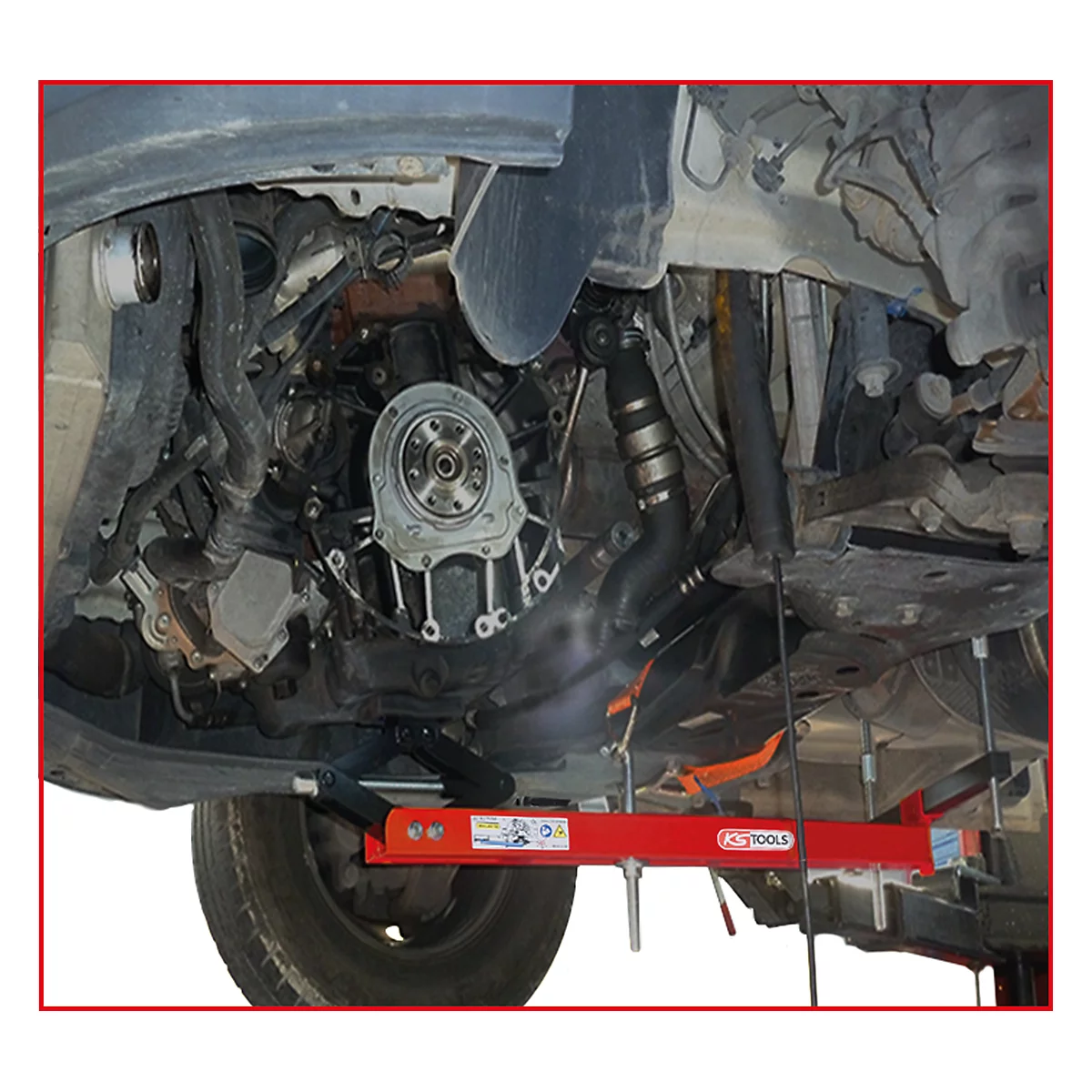 Unteransicht eines Autos. Eine Hebebühne hält den Motor. Teile des Motors sind zu sehen, darunter ein Rad und Rohre.